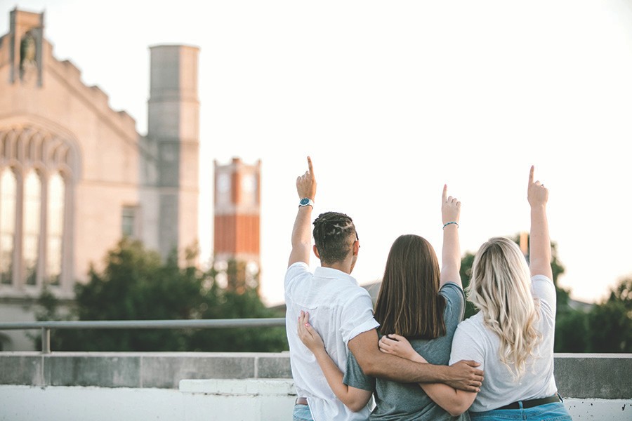 Three students raising symbolic one finger in the air while embracing one another side by side on a rooftop facing Sutton Hall with the Bizzell Clocktower in the distance.