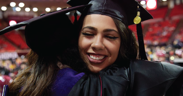 Two students in graduation regalia hugging.