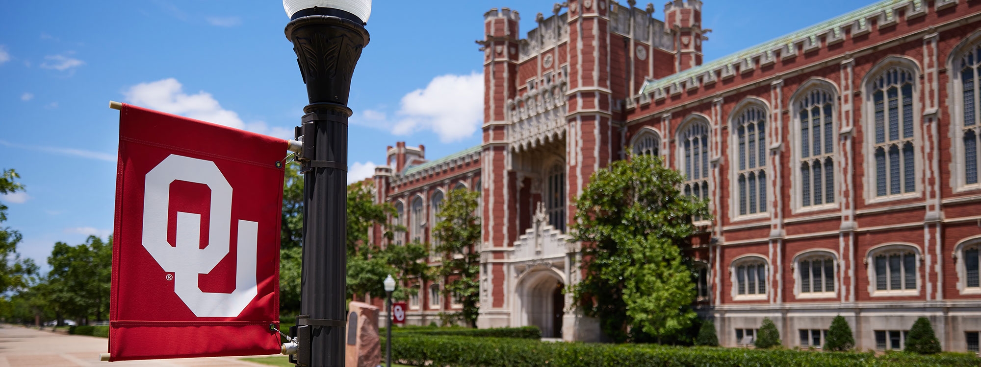 Bizzell Memorial Library with a blue sky in the background and a lamppost with OU flag in the foreground.