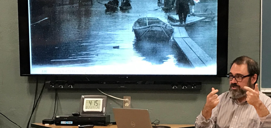 Photo of Professor Soppelsa discussing an early 20th century French postcard image on the large screen to his right