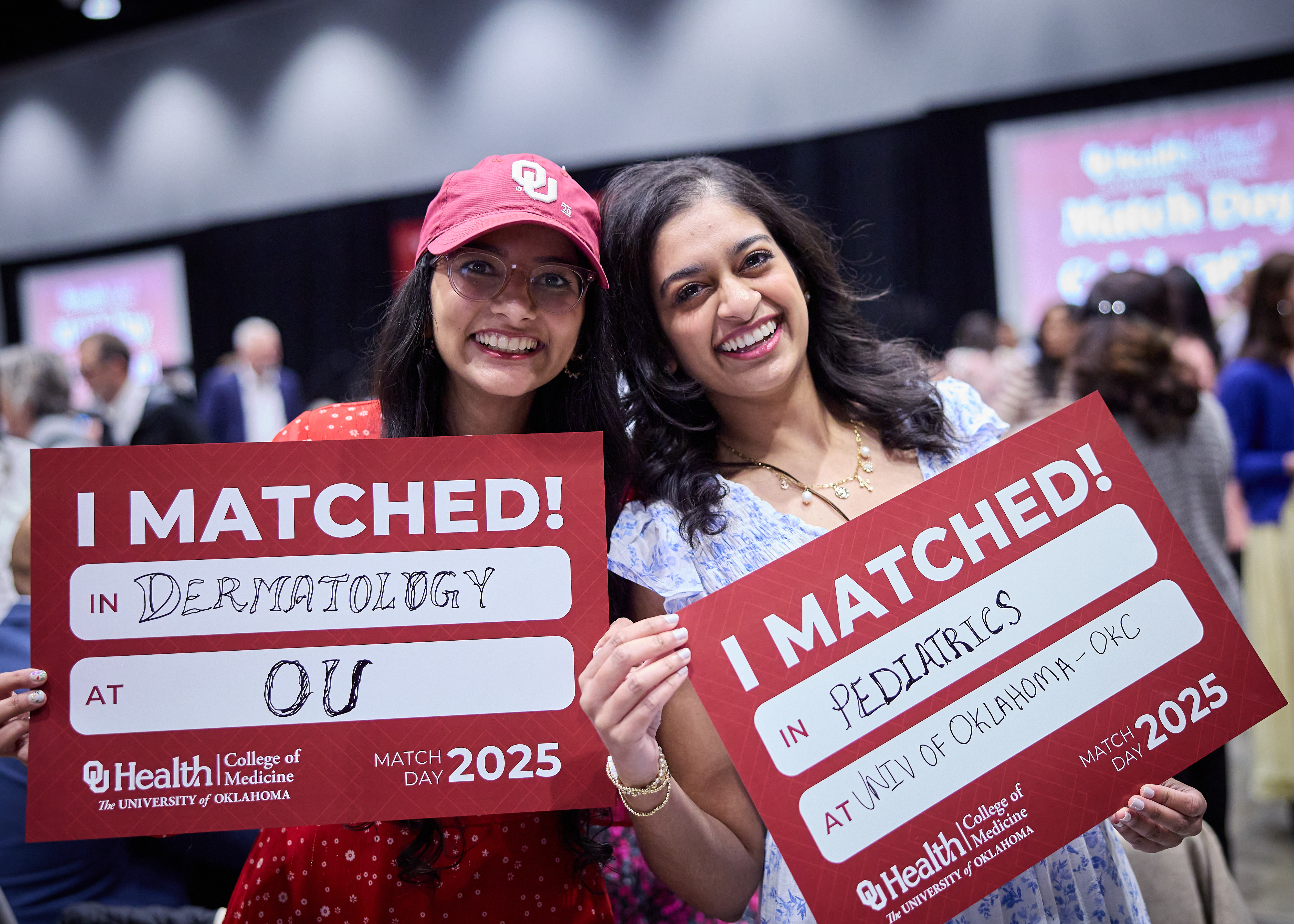 Two medical students hold up signs showing which department and at OU they matched into.