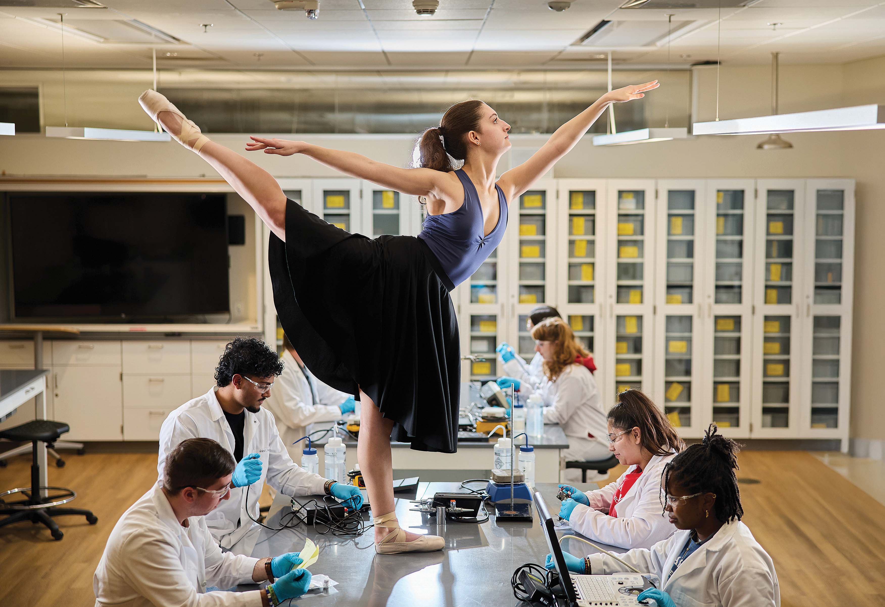 A ballerina lifting holding a pose on a laboratory table while students work on science.