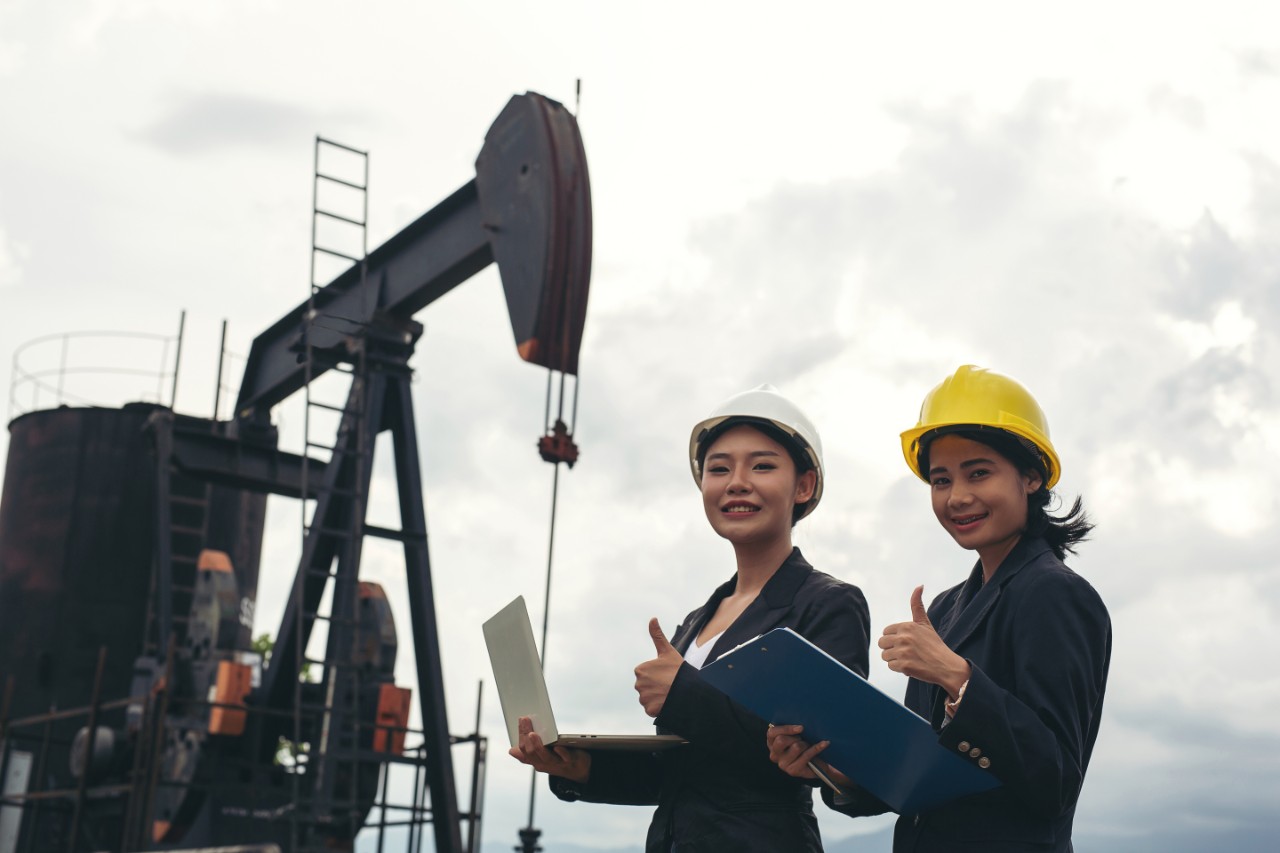 Two female engineers stand beside working oil pumps with a white sky background. 2-interns-next-to-pump-in-the-field