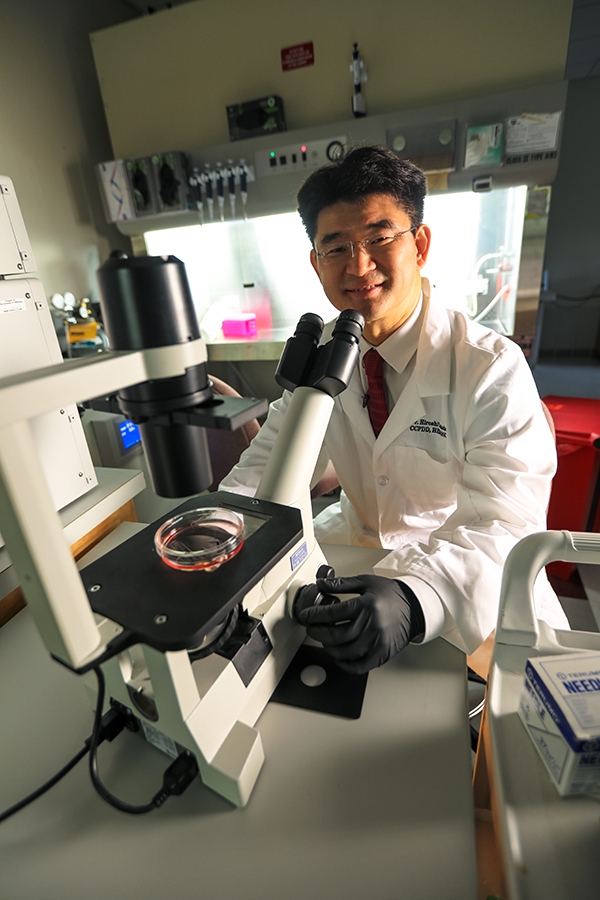 Hiroshi Yamada, Ph.D., sitting at a microscope in a lab smiling.