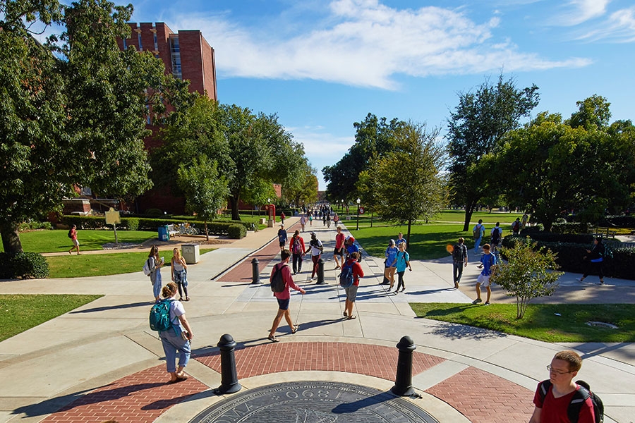 Students walking on the OU Scholars Walk, with trees, buildings, and a blue sky in the background.
