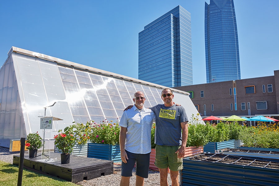Two men stand in front of a greenhouse.