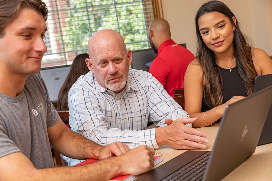 A professor sitting at a table with two students using laptops. 