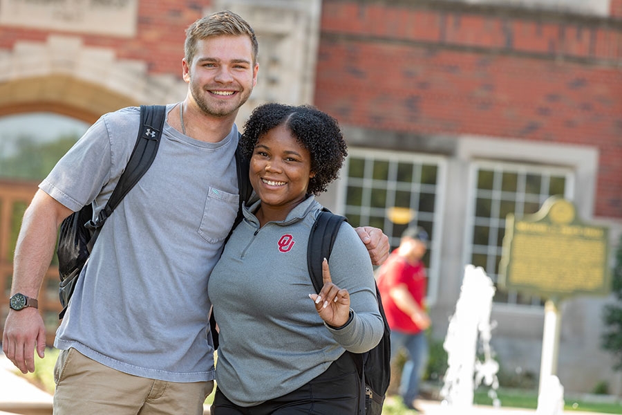 Two students standing together outside Price Hall. One is giving the "Only One" hand gesture.