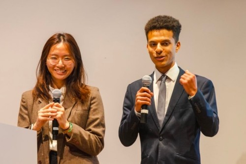 Two students, holding microphones, stand near a podium while giving final presentations