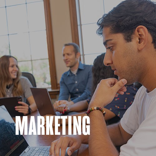 A student working on a laptop with other students nearby. Text over photo reads: Marketing.