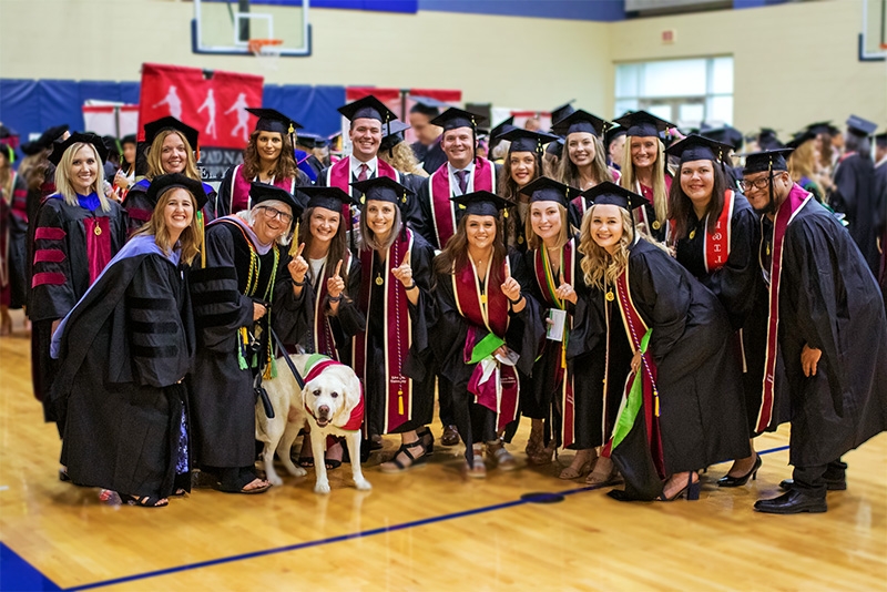 A group of OU-Tulsa graduates stand together to take a photo in their cap and gown.