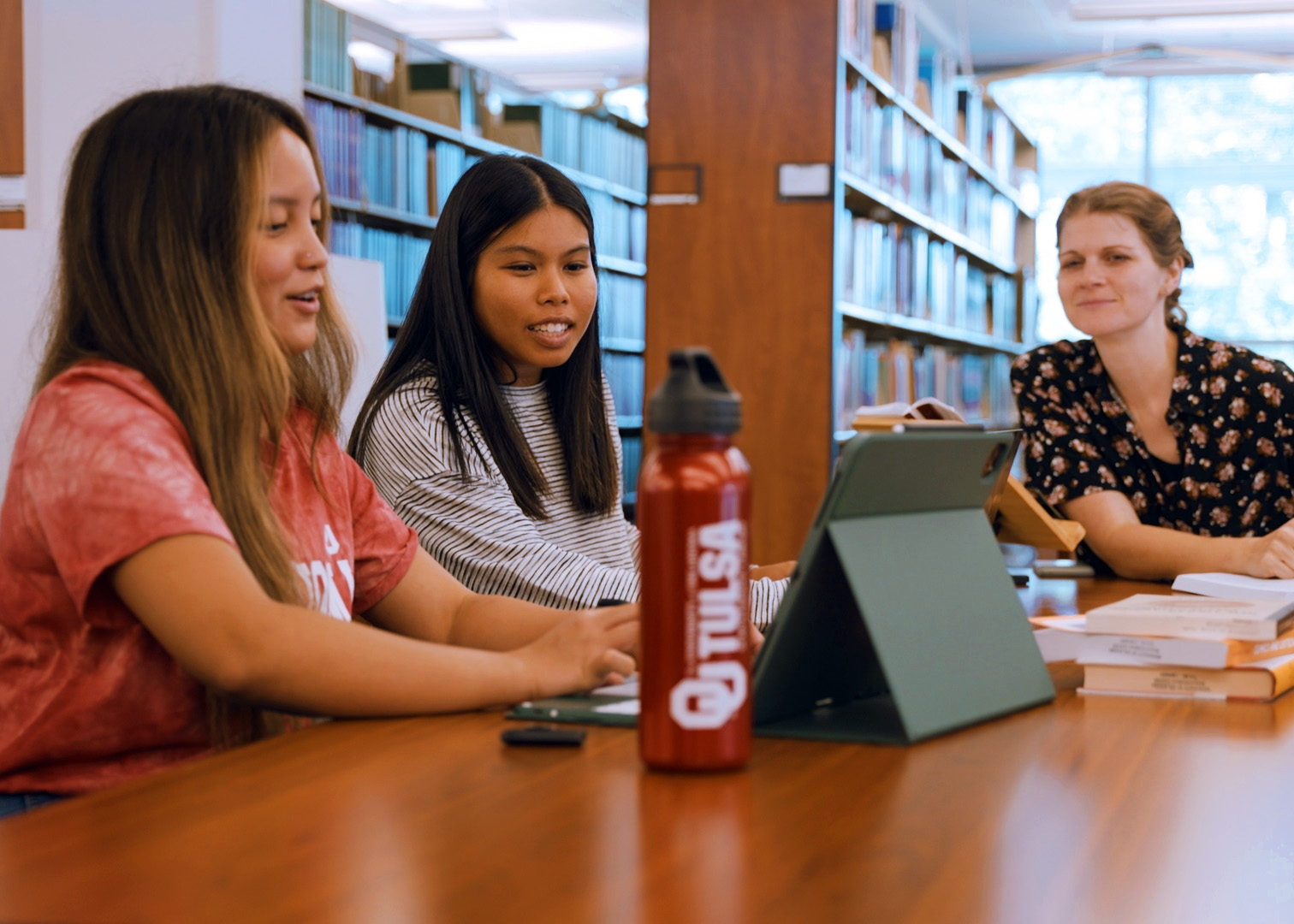 Three students sit at a table in a library setting, looking over work on a tablet computer